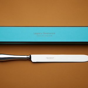 Puede incluir: Un cuchillo para pastel de plata con una caja turquesa. La caja tiene el texto "Legacy Silverware".