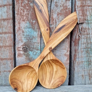 May include: Two wooden spoons with long handles and rounded bowls. The spoons are made of light brown wood with a visible grain pattern. The spoons are arranged in a crossed position against a weathered wooden background.
