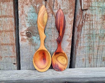 May include: Two handcrafted wooden spoons with distinct grain patterns. The left spoon features a light golden-brown hue with swirling wood grain, while the right spoon displays a deeper reddish-brown color with prominent, dark, circular grain rings.