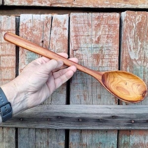 May include: A long-handled wooden spoon with a smooth, polished finish and a visible natural grain pattern. The spoon features a deep, oval-shaped bowl and a sturdy, straight handle, showcasing warm brown and golden tones throughout the wood.