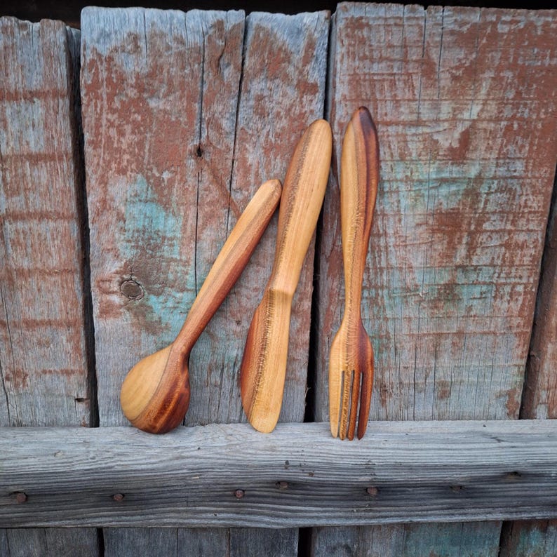 May include: A wooden spoon, knife, and fork set. The utensils are made of light brown wood with darker brown grain patterns. The spoon has a rounded bowl, the knife has a flat blade, and the fork has three tines. The set is displayed against a weathered wooden background.