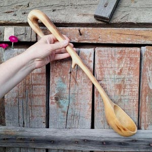 May include: A long-handled wooden ladle carved from a single piece of light-colored wood. The handle features a natural, curved branch-like shape with a distinct knot and a loop at the top, leading down to a smooth, oval-shaped bowl at the end.