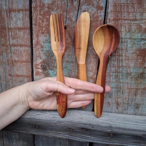 May include: A wooden fork, knife, and spoon set. The utensils are made of light and dark brown wood. The fork has four tines, the knife has a rounded tip, and the spoon has a deep bowl. The set is displayed against a weathered wooden background.