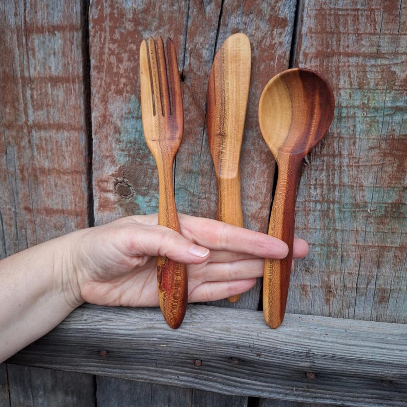 May include: A wooden fork, knife, and spoon set. The utensils are made of light and dark brown wood. The fork has four tines, the knife has a rounded tip, and the spoon has a deep bowl. The set is displayed against a weathered wooden background.