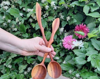 May include: Two handcrafted wooden spoons with a natural wood grain finish. The spoons are held against a backdrop of green foliage, white and pink flowers. The spoons have a curved handle and a rounded bowl.
