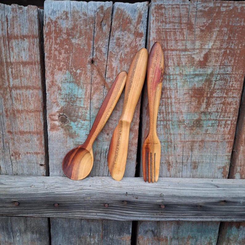 May include: A wooden spoon, knife, and fork set. The spoon has a rounded bowl and long handle. The knife has a flat blade and a handle. The fork has three tines. The utensils are made of light brown wood with reddish-brown accents.