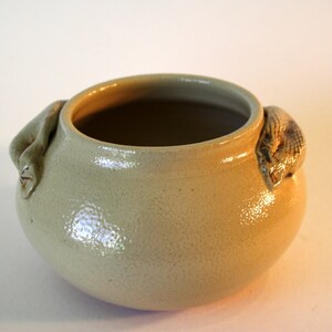 Könnte beinhalten: Ein kleiner, cremefarbener Keramiktopf mit zwei geformten Griffen. Die Griffe sind wie eingerollte Schlangen geformt.