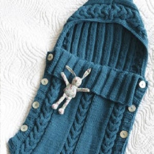 Op de afbeelding: Een baby slaapzak van tealblauwe gebreide stof met een capuchon en knopen. De slaapzak heeft een kabelbreipatroon en een klein knuffelkonijn zit erin.