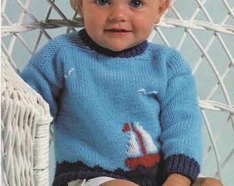 May include: A blue knitted sweater with a white sailboat design for a baby. The sweater has a navy blue trim around the neck and sleeves. The baby is wearing white shorts and sitting in a white wicker chair.
