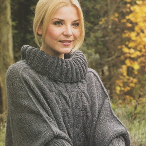 Puede incluir: Un poncho de punto gris con cuello alto de tortuga y patrón de punto de cable. El poncho lo lleva una persona que sostiene una herramienta de jardinería.