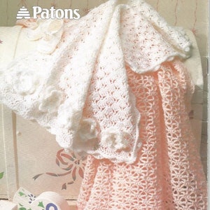 May include: Two crocheted baby blankets, one white and one pink, are draped over a white wooden box. The box has a floral design painted on it. The blankets are made with a delicate, lacy stitch. The label "Patons" is visible on the box.