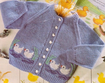 May include: A light blue knitted baby cardigan with white buttons and embroidered ducks on the front. The cardigan is laying on a white wooden surface with a children's book in the background.