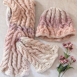 May include: A knitted hat and scarf set in a pink and cream ombre colorway. The hat is a slouchy beanie style and the scarf is a long, rectangular shape. Both pieces are made with a cable knit stitch.