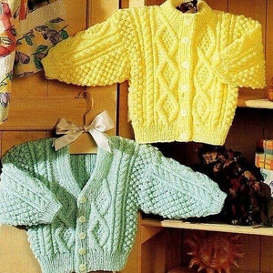 Puede incluir: Dos cárdigans de punto para bebé, uno amarillo y otro azul claro, con un patrón de punto de cable y cierres de botón. Los cárdigans están colgados en un estante de madera.