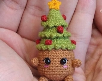 以下が含まれることがあります： 茶色の顔と大きな目を持つ、小さな手編みのクリスマスツリーのオーナメント。木は緑色で、赤いオーナメントと黄色い星が頂上にあります。