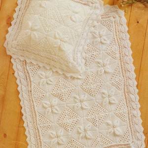 Può includere: Set di coperta e cuscino per neonati, lavorati all'uncinetto a mano, color crema. La coperta presenta un motivo floreale all'interno di forme a diamante, con un bordo smerlato. Il cuscino ha un design floreale simile. Il set è esposto su una superficie di legno.