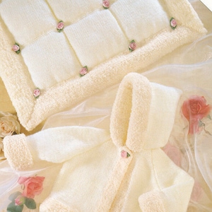 Pode incluir: Uma jaqueta de tricô branca para bebê com capuz e fecho de botão. A jaqueta tem uma bainha branca felpuda ao redor das bordas e um enfeite de flor rosa na frente. A jaqueta está colocada sobre um fundo de tecido branco com um travesseiro de tricô branco com enfeites de flores rosas no fundo.