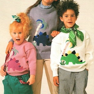 Pode incluir: Três crianças vestindo suéteres de tricô com desenhos de dinossauros e palmeiras. Os suéteres são rosa, roxo e branco. As crianças estão sorrindo e olhando para a câmera.