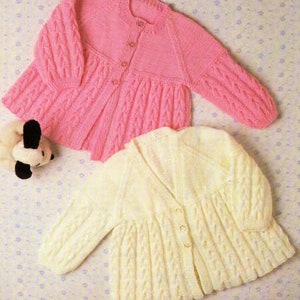 May include: Two knitted baby cardigans, one pink and one cream, with a cable knit pattern and button closures.