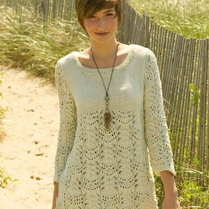 May include: A woman wearing a light beige knitted sweater with a delicate lace pattern. The sweater has a round neckline and long sleeves. She is wearing a brown pendant necklace with a tassel.