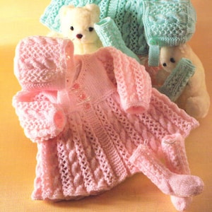 May include: A pink knitted baby dress with a matching bonnet and booties. The dress has a cable knit pattern and a button closure at the front. The bonnet and booties are also cable knit.
