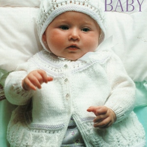 May include: A white knitted baby cardigan with a button closure and a matching white knitted hat. The cardigan has a delicate lace pattern and a scalloped edge. The hat has a flower design.