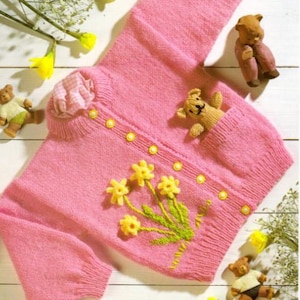 May include: A pink knitted cardigan with a button closure and a pocket on the front. The cardigan has a floral design embroidered on the front and is decorated with yellow buttons. The text "See back page for detail of toy" is printed on the top right corner of the image.