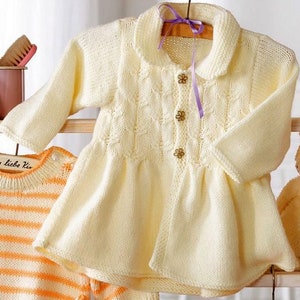 May include: Cream-colored knitted baby cardigan with a Peter Pan collar and three flower-shaped buttons. The long-sleeved sweater has a flared skirt and is hanging on a wooden hanger. A purple ribbon is tied at the top.