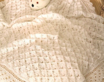 Op de afbeelding: Een wit gebreid babydekentje met een delicate kantzoom. Een bruin konijn en een witte muis zitten op het dekentje.
