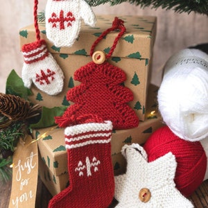 May include: A set of knitted Christmas decorations, including a stocking, a star, a mitten, and a Christmas tree. The decorations are red and white with a Scandinavian design. The decorations are displayed on a brown box with a gift tag that says "Just for you".