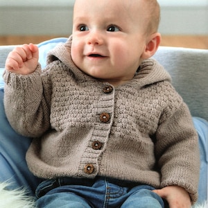 Könnte beinhalten: Ein Baby trägt einen braunen Strickpullover mit Kapuze und Holzknöpfen. Der Pullover hat ein strukturiertes Muster und das Baby lächelt.
