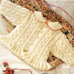 May include: Cream-colored knitted baby cardigan with a Peter Pan collar trimmed in red. The sweater features pearl-like buttons and a ruffled hem. The sleeves and body have textured cable and wave patterns. A delicate, handmade garment.