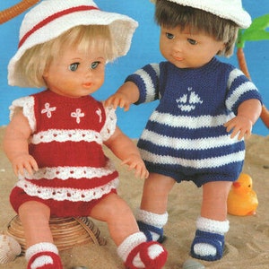 Op de afbeelding: Twee poppen in gebreide outfits en hoeden. Eén pop draagt een rood-wit gestreepte romper met bijpassende hoed. De andere pop heeft een blauw-wit gestreepte outfit met een witte hoed aan. Ze staan op een zanderige ondergrond.
