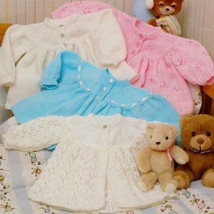 May include: Four knitted baby cardigans in white, pink, blue, and cream. The cardigans are laid out on a white and blue patterned fabric with two teddy bears.