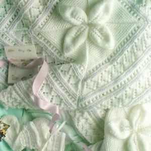 May include: A white knitted baby blanket with a delicate floral design and matching booties. The blanket features a large flower in the centre and a lacy border.