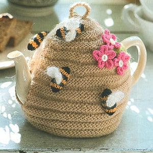 Könnte beinhalten: Eine braune gestrickte Teekannenhülle mit einem Bienenstockmuster. Die Hülle ist mit gestickten Bienen und rosa Blumen verziert. Die Teekanne ist weiß mit einem braunen Henkel.