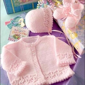 May include: A pink knitted baby cardigan with a button closure, matching hat, and booties. The set is made of soft yarn and features delicate lace detailing.