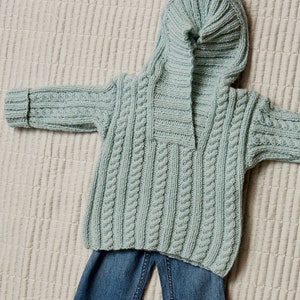 May include: A light blue knitted sweater with a hood and a cable knit pattern. The sweater is laid out on a white textured surface with a pair of blue jeans underneath.
