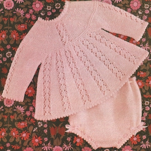 May include: A pink knitted baby outfit with a lacy top and matching trousers. The outfit is laid out on a floral fabric background.