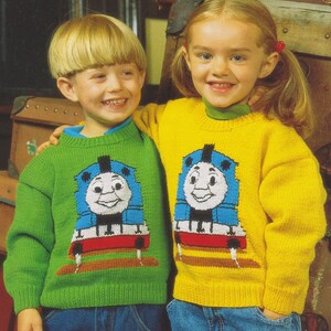 Może przedstawiać: Dwoje dzieci w zielonych i żółtych swetrach z motywami Thomasa the Tank Engine. Swetry są dziane i przedstawiają niebieski i czerwony pociąg z uśmiechniętą twarzą.