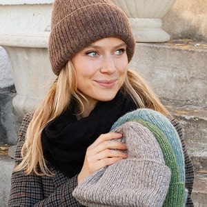 Pode incluir: Uma mulher está usando um gorro de tricô marrom e segurando três gorros de tricô em suas mãos. Os gorros são em tons de marrom, verde e azul.