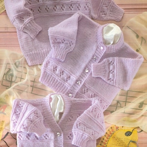 May include: Three lavender knitted baby cardigans with button closures. The cardigans have a delicate lace pattern and are made with soft yarn.