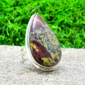 May include: A silver ring with a teardrop-shaped red, green, and yellow gemstone. The ring is set on a gray rock with a green moss background.