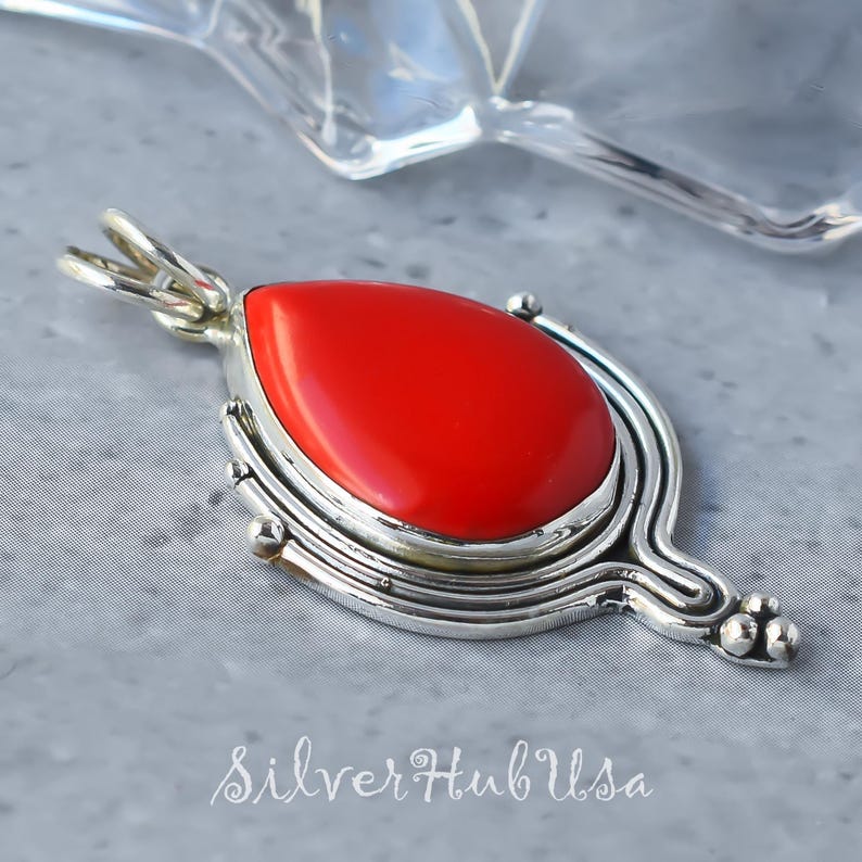 Puede incluir: Un colgante de plata con una piedra roja brillante en forma de l&aacute;grima engastada en un bisel de plata. El colgante tiene un dise&ntilde;o detallado con un patr&oacute;n de espiral y peque&ntilde;as cuentas.