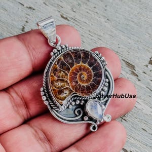 May include: A silver pendant with a polished ammonite fossil, displaying a spiral pattern in brown and gold hues. The pendant incorporates a small, pear-shaped, iridescent stone and is suspended from a silver bail.
