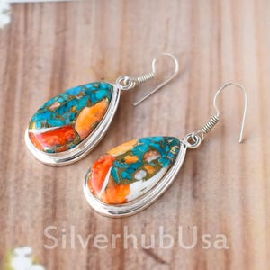 May include: A pair of silver dangle earrings with teardrop-shaped turquoise, coral, and white stone inlays. The earrings have a silver hook fastening.