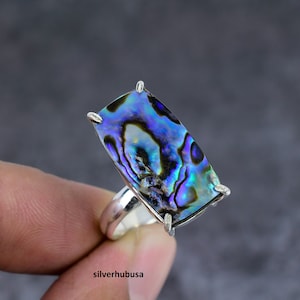 Puede incluir: Un anillo de plata con una gran piedra rectangular de concha de abulón. La concha tiene colores iridiscentes azules, verdes y morados.