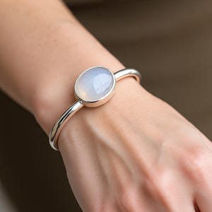 May include: A silver bangle bracelet featuring an oval, pale blue gemstone. The gemstone is set in a silver bezel and is the focal point of the bracelet. The bracelet has a simple, elegant design.