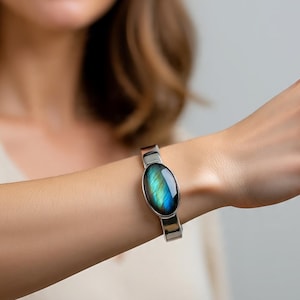 Könnte beinhalten: Ein silberfarbenes Armband mit einem ovalen Labradoritstein mit schillernden blauen und grünen Farbtönen. Der Stein ist in einen polierten Metallrahmen gefasst, und das Armband hat ein schlichtes, modernes Design. Das Armband wird am Handgelenk getragen.