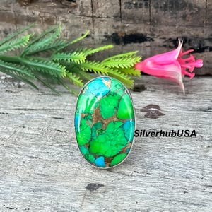 Puede incluir: Anillo ovalado de plata con una piedra en tonos verdes, turquesas y dorados. La piedra tiene una superficie texturizada. El anillo se muestra sobre una superficie de madera, con vegetación verde y una flor rosa de fondo.
